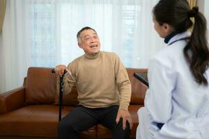 Doctor examines and treats an elderly man seated on a sofa with a cane who is suffering from knee joint pain. photo