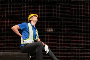 retrato de un masculino trabajador vistiendo un la seguridad chaleco y casco sentado en un aceros paleta debido a espalda dolor desde trabajando en un fábrica levantamiento pesado cosas. foto