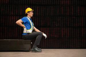 Portrait of a male worker wearing a safety vest and helmet sitting on a steels pallet due to back pain from working in a factory lifting heavy things. photo