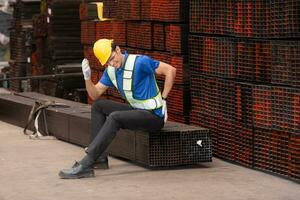 retrato de un masculino trabajador vistiendo un la seguridad chaleco y casco sentado en un aceros paleta debido a espalda dolor desde trabajando en un fábrica levantamiento pesado cosas. foto