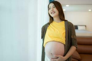 Beautiful pregnant woman standing near window at home and holding her belly photo