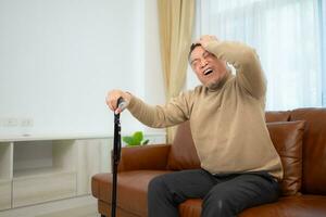 With headache, a senior guy sits on a sofa with a walking stick in his living room at home. photo