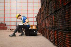 Portrait of a male worker wearing a safety vest and helmet sitting on a steels pallet due to back pain from working in a factory lifting heavy things. photo