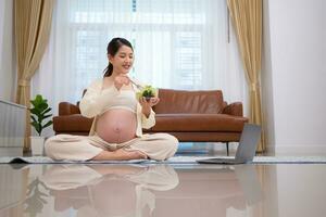 embarazada mujer come ensalada como un bocadillo mientras sentado en el piso de su hogar. foto