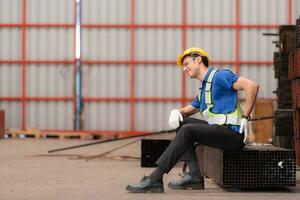 retrato de un masculino trabajador vistiendo un la seguridad chaleco y casco sentado en un aceros paleta debido a espalda dolor desde trabajando en un fábrica levantamiento pesado cosas. foto