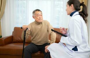 Doctor examines and treats an elderly man seated on a sofa with a cane who is suffering from knee joint pain. photo