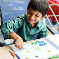 Smart Indian little boy perform thumb painting with different colourful water colour kit during the summer vacations, Cute Indian Kid doing colourful thumb painting drawing on wooden table photo