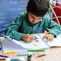 Smart Indian little boy perform thumb painting with different colourful water colour kit during the summer vacations, Cute Indian Kid doing colourful thumb painting drawing on wooden table photo