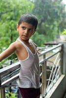 pequeño niño jugando en verano lluvia en casa balcón, indio inteligente chico jugando con lluvia gotas durante monzón lluvioso estación, niño jugando en lluvia foto