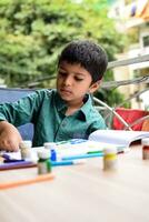 Smart Indian little boy perform thumb painting with different colourful water colour kit during the summer vacations, Cute Indian Kid doing colourful thumb painting drawing on wooden table photo