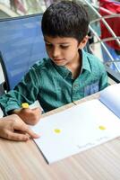 Smart Indian little boy perform thumb painting with different colourful water colour kit during the summer vacations, Cute Indian Kid doing colourful thumb painting drawing on wooden table photo