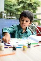 Smart Indian little boy perform thumb painting with different colourful water colour kit during the summer vacations, Cute Indian Kid doing colourful thumb painting drawing on wooden table photo