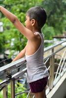 pequeño niño jugando en verano lluvia en casa balcón, indio inteligente chico jugando con lluvia gotas durante monzón lluvioso estación, niño jugando en lluvia foto