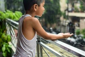 Little kid playing in summer rain in house balcony, Indian smart boy playing with rain drops during monsoon rainy season, kid playing in rain photo