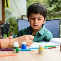 Smart Indian little boy perform thumb painting with different colourful water colour kit during the summer vacations, Cute Indian Kid doing colourful thumb painting drawing on wooden table photo