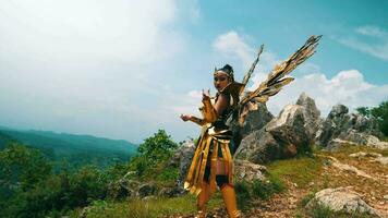 a female warrior in a golden costume and wings standing on a rocky mountain against video