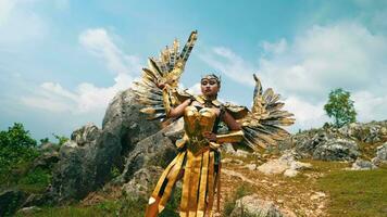 a female warrior in a golden costume and wings standing on a rocky mountain against video