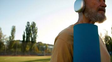 Alten kaukasisch Mann im Kopfhörer suchen beim Armbanduhr Sport Zeit Morgen Stadion Übung Athlet Ausbildung Matte physisch Aktivität Lebensstil Gesundheit Wohlbefinden Stadt draußen motiviert Fitness Ausrüstung video