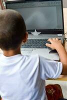 pequeño chico sentado a mesa utilizando ordenador portátil para en línea clase en grado 1, niño estudiando en ordenador portátil desde hogar para distancia aprendizaje en línea educación, colegio chico niños estilo de vida concepto foto