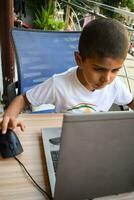 pequeño chico sentado a mesa utilizando ordenador portátil para en línea clase en grado 1, niño estudiando en ordenador portátil desde hogar para distancia aprendizaje en línea educación, colegio chico niños estilo de vida concepto foto