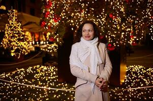 maravilloso mujer disfruta el caminar en Navidad justo, admira hermosa ciudad calle decorado con festivo guirnaldas a noche foto