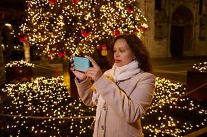 hermosa Rizado peludo morena mujer fotografiando el Navidad recinto ferial en el ciudad cuadrado a noche foto