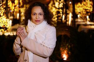 Attractive young woman looking confidently at camera standing against Christmas lights backdrop at funfair at night time photo