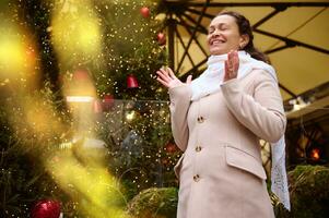 Photo with bokeh on foreground while a charming happy woman smiling broadly, making cherished wish at Christmas funfair