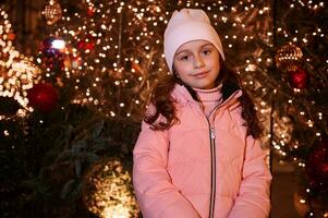 Beautiful little kid girl smiling at camera, standing against Christmas tree at fairground or traditional family market photo