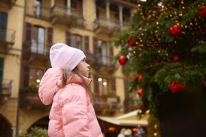 pequeño niña hace querido desear, en pie en el calle, admirativo un hermosa Navidad árbol, decorado con iluminaciones foto