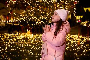 linda niño niña con manos ahuecado, haciendo deseo para Navidad, en contra un calle iluminado por Navidad luces a un justa foto