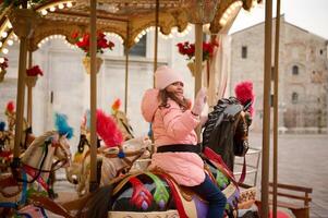 Happy child girl smiles and waves hello with hand while having fun on a merry go round carousel at Christmas funfair. photo