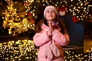 pequeño niña con ahuecado manos, hace querido deseo para Navidad, en pie a recinto ferial, disfrutando festivo atmósfera foto
