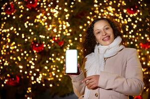 Cheerful Latina woman showing mobile phone with white mockup screen, for ads or application, against lighted Xmas tree photo