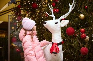 Caucasian kid girl standing by Christmas deer against festive Xmas tree, decorated with garlands and balls. December 25 photo