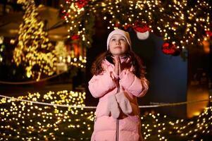 adorable niño niña participación manos palmas juntos, haciendo querido desear, en pie en contra un Navidad árbol a parque de atracciones foto