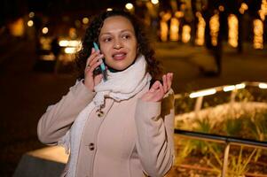 Multi ethnic young adult woman smiles while talks on mobile phone, standing in the illuminated city street by night photo