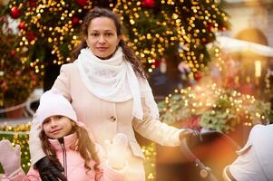 hermosa madre con bebé cochecito y su linda hija mirando a cámara, en pie en contra un Navidad árbol al aire libre foto