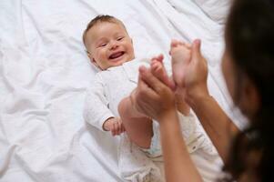 hermosa recién nacido bebé sonrisas mirando a su amoroso madre, mientras ella golpes y Besos su pequeño pies y minúsculo dedos de los pies foto