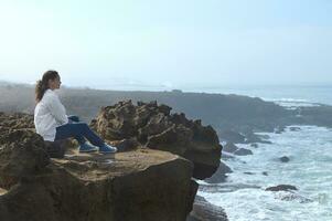 Relaxed multi ethnic woman sitting alone on a cliff and admires the mesmerizing view of the Atlantic Ocean. photo