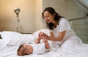 contento encantador cariñoso joven madre cuidando de su recién nacido bebé chico, acariciando y besos su pequeño pies y dedos de los pies foto