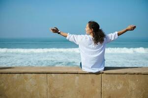 Happy curly haired woman outstretches her arms to the sides, sitting by Atlantic ocean and enjoying mesmerizing nature photo