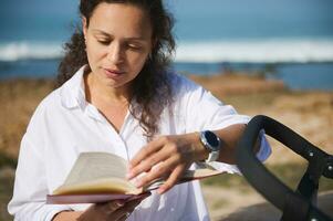 hermosa mujer sentado cerca su bebé paseante, hojeando mediante paginas mientras leyendo novela en el playa. digital desintoxicación foto