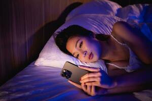 asiático mujer utilizando móvil teléfono teléfono inteligente tendido en el cama en el dormitorio. soñoliento exhausto, lata no dormir. insomnio, adiccion concepto. mujer desplazamiento social redes en móvil oscuro dormitorio. foto