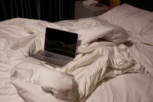 Laptop on unmade bed with messy sheets and sleeping pillows photo