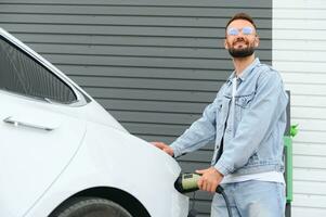 hermosa joven elegante hombre es con eléctrico coche a tiempo de día cargando el vehículo foto