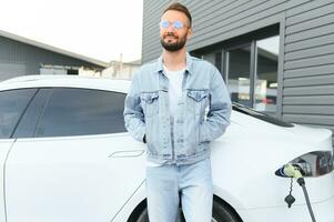 Standing and waiting for charge to finish. Young stylish man is with electric car at daytime photo