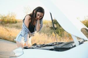 Beautiful slim girl in shirt and shorts looks in open car hood on a road photo