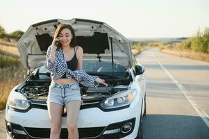 atractivo Delgado joven niña en verano pantalones cortos y camisa refacción un roto coche. un hermosa mujer soportes cerca elevado coche capucha. foto