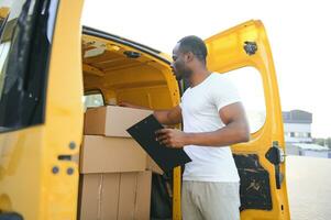 african american Worker unloading boxes from van outdoors. Moving service concept. Male employee of moving company makes inventory of packed boxes and writes data to clipboard. photo